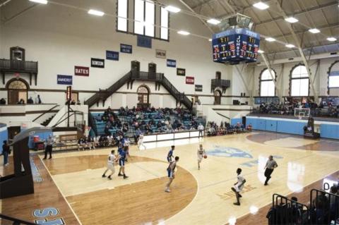 The Medford and Somerville High Schools' boys basketball teams play each other in a sunlit Tufts-branded gymnasium as stands full of spectators look on.