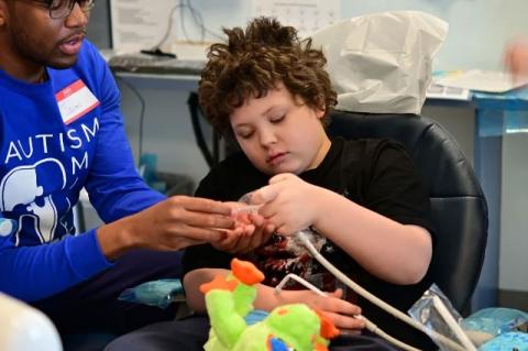 A child in a dental chair handling some dental accessories handed to him by a dental student. The Tufts Autism Smiles program helps children on the autism spectrum become comfortable with dental offices in preparation for a visit to the dentist.