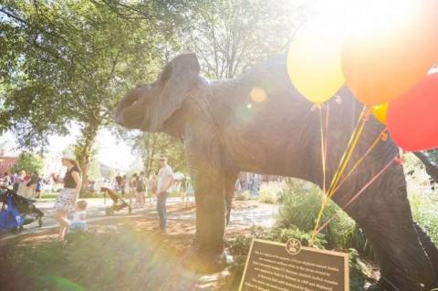 A statue of Jumbo the elephant in the sunlight is in front of a green space filled with community members celebrating Community Day