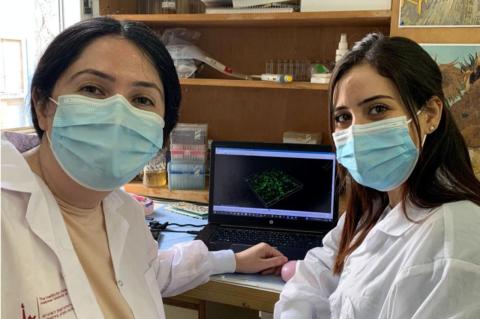 Two women in lab coats wearing blue surgical masks, in front of a computer monitor. A program pairs Palestinian and Israeli graduate students to work together toward their degrees.