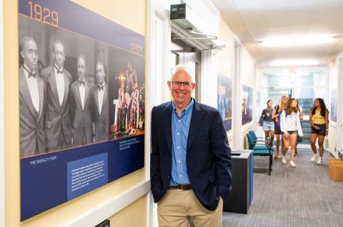 James Rice stands in front of a photo display at the Jumbo Generations exhibition in East Hall, with students walking behind him. A new exhibition of photographs from the 1870s to the present show the ways that Tufts campus life has evolved