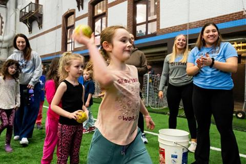 A young girl who attended the National Girls and Women in Sports Day event at Tufts University on Saturday, February 3, throwing a softball while members of the Tufts women&#039;s softball team cheer her on in the background.