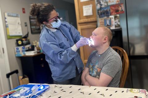 A dental hygienist examines a young man sitting at a kitchen table.