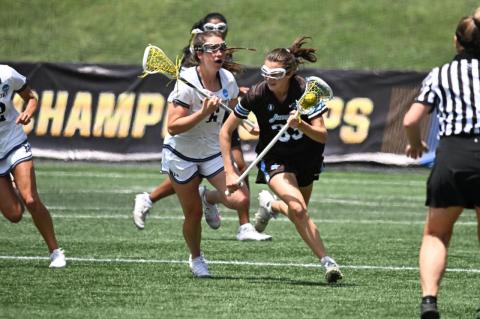 A women’s lacrosse player in brown with the ball runs past a player in white, as a referee looks on
