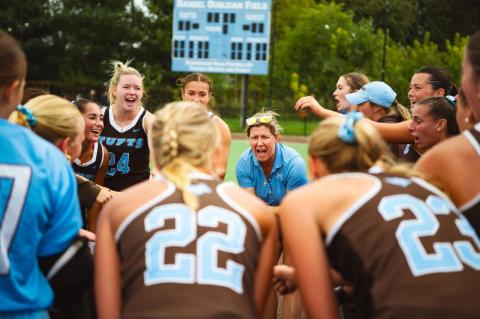 A group of women athletes in brown and blue jerseys stand in a circle and cheer