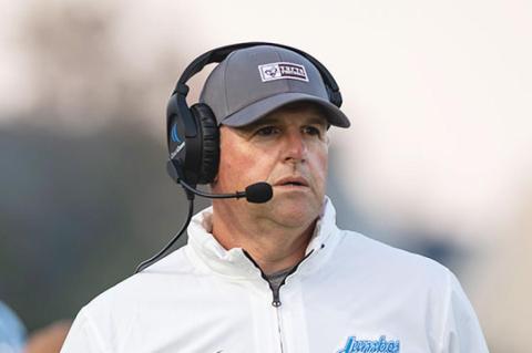 A man wearing a white polo and baseball cap talks into a headset while standing on a football field