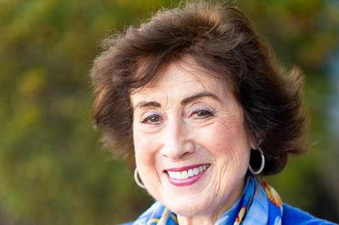 Person wearing a bright blue top and a colorful patterned scarf with shades of blue, yellow, and red, standing outdoors with greenery in the background.