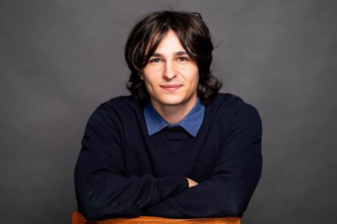 Shai Tratt sits leaning against the back of a chair, seen against a gray backdrop