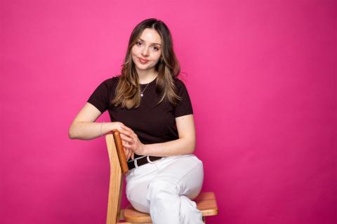 A woman sits in a chair in front of a bright pink background. 