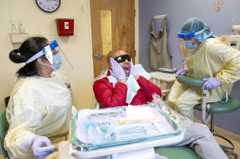 Man in a red shirt opens his mouth wide while in the dental chair, with two dental workers at his side.