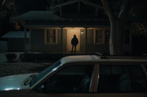 A woman stands in front of a house across from a parked car